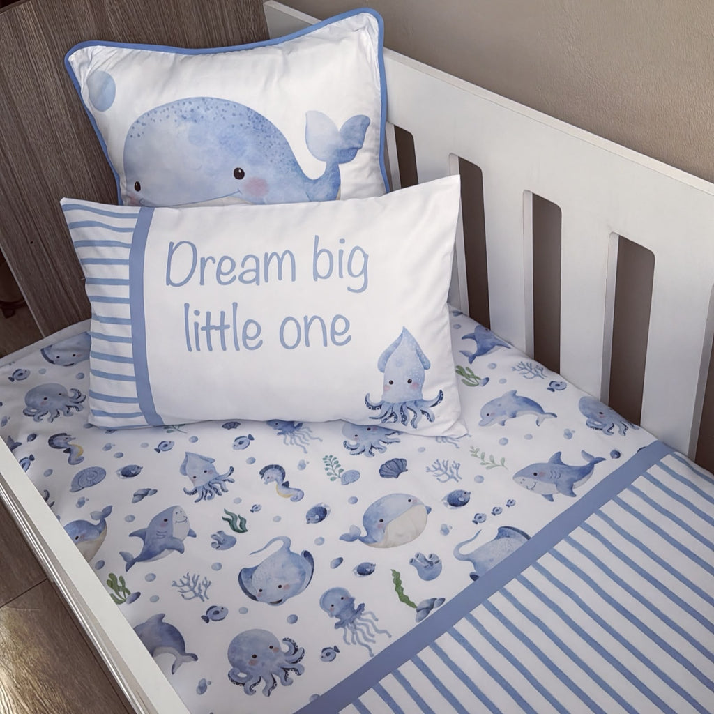 A blue and white baby bedding set styled in a white wooden crib, featuring a duvet cover with a playful sea-creature print, a striped pillow, and a whale-print pillow.