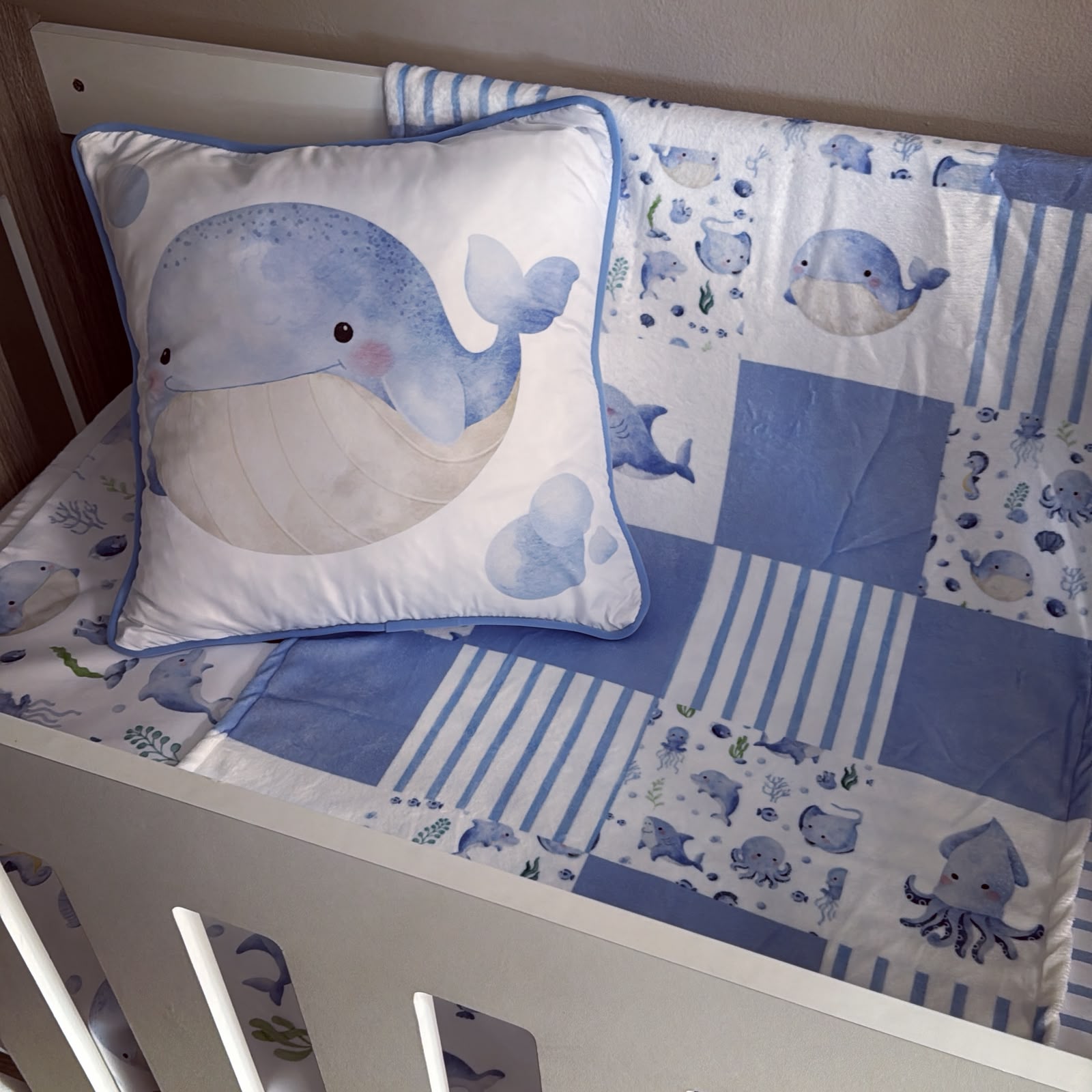 A blue and white whale-themed crib bedding set including a patchwork quilt, matching crib bumper, and a decorative whale pillow, shown inside a white wooden crib.