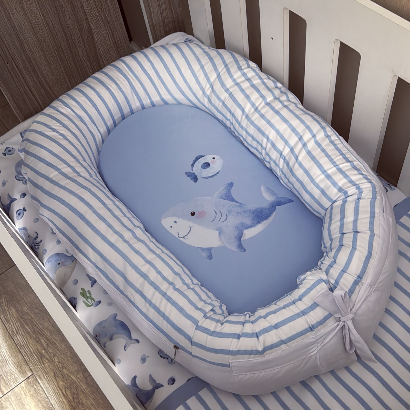 A baby bassinet cushion set with a light blue and white striped fabric border and a matching blue insert featuring a shark and whale illustration, shown inside a white crib.
