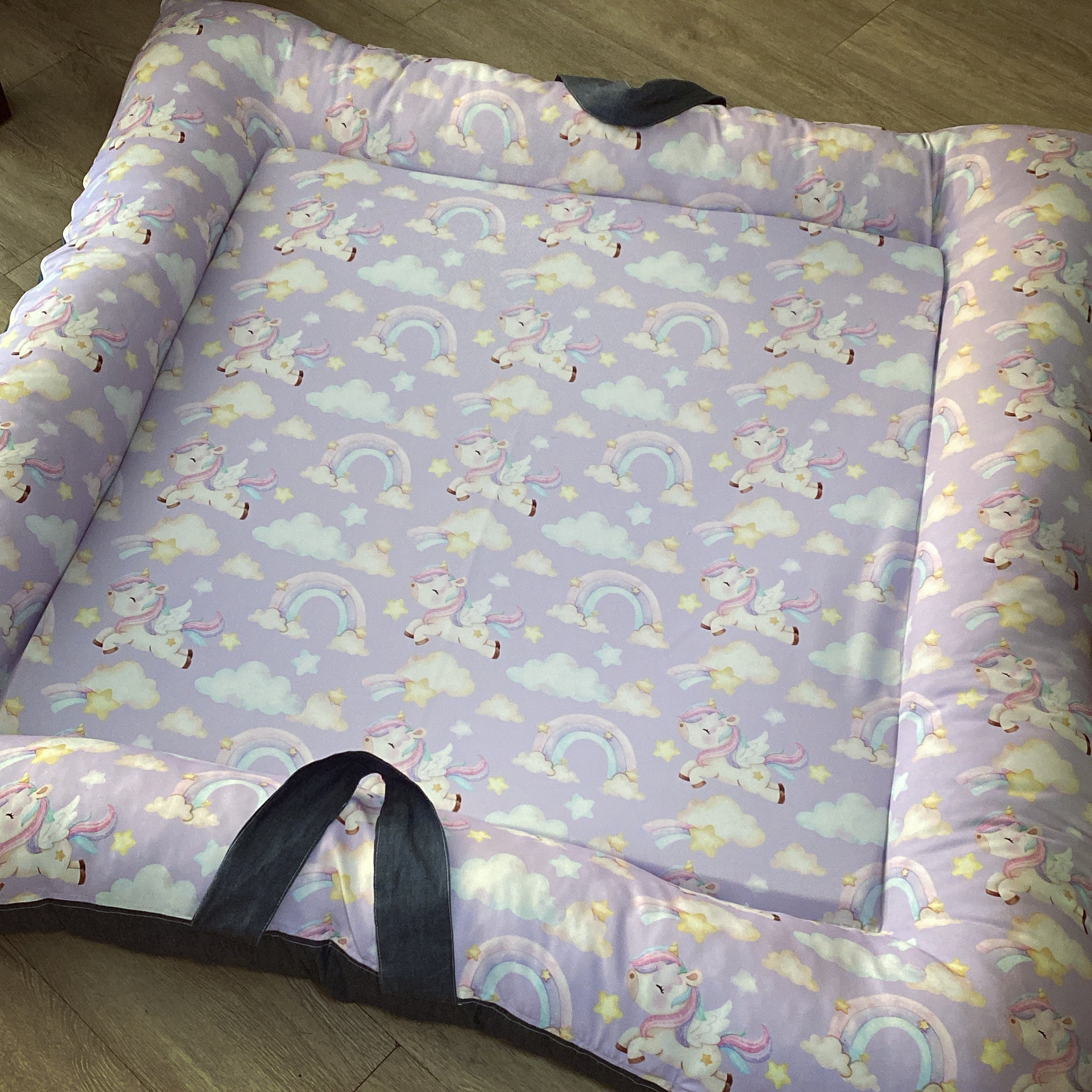 A multicolored baby play doughnut with a unicorn and rainbow pattern on a purple background, with side handles.