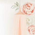 A pink hanging tent canopy with floral designs, set up in a room with a white background. It is positioned above a cream-colored feeding chair with a rose gold accent.