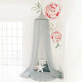 A grey hanging tent canopy with floral netting patterns set up in a room with two large pink artificial roses and a small cushion underneath.