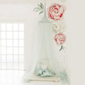 A moss green hanging tent canopy with a hoop diameter of 45cm, adorned with artificial pink roses and green foliage, set up in a room with a white cushion and a small star-shaped pillow.