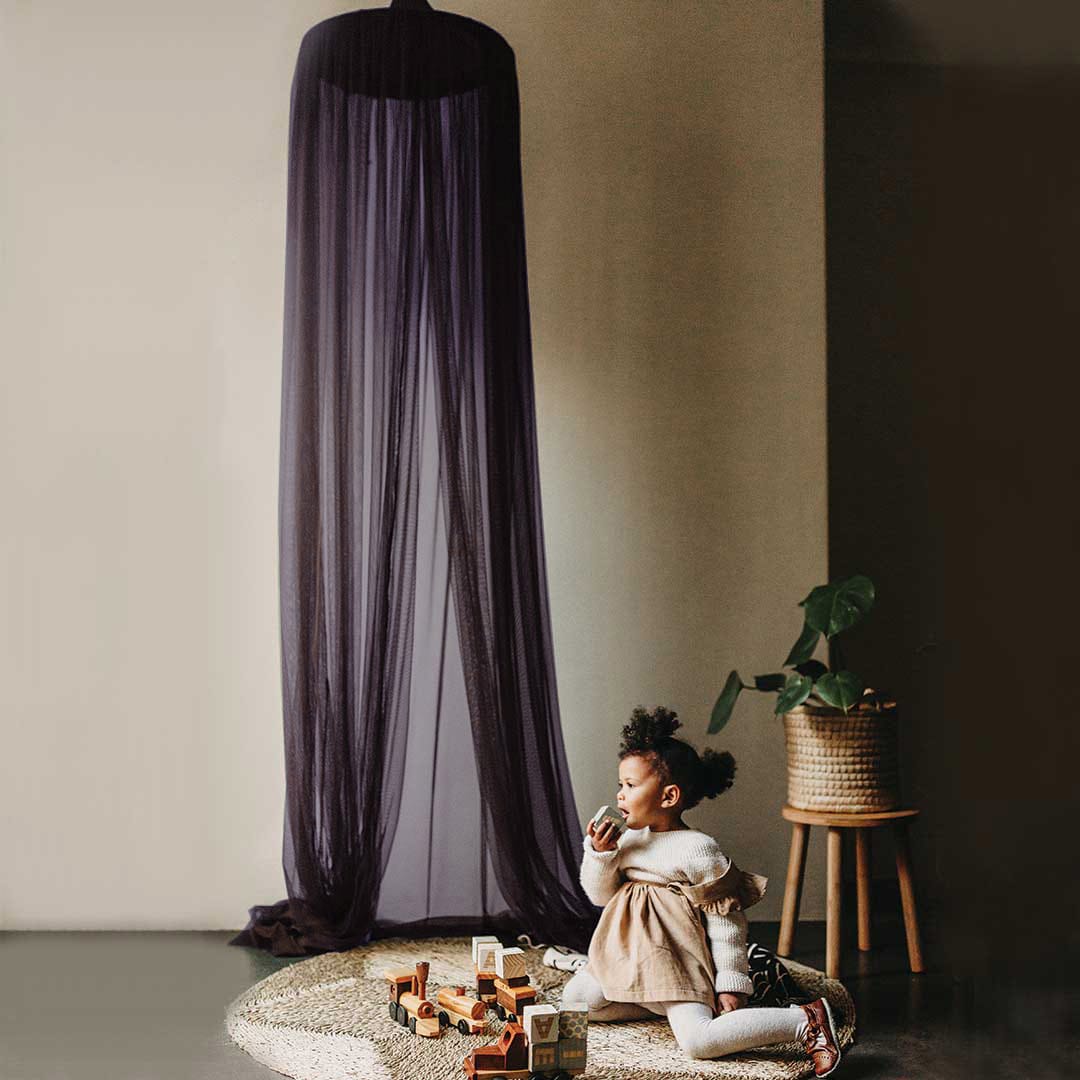 A plum-colored hanging tent canopy set up on a wall, with a child sitting beneath it looking at toys on the ground.