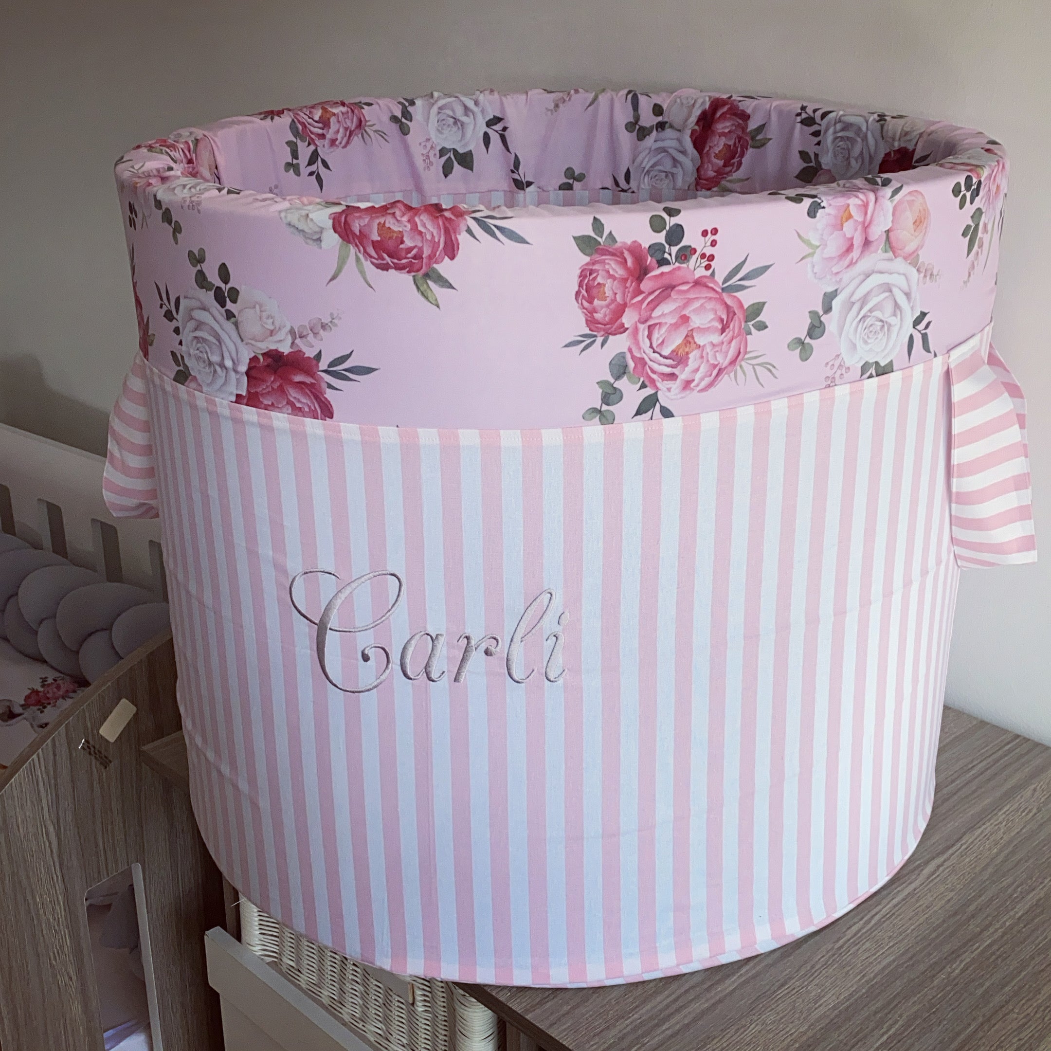 A large laundry barrel with a pink and white striped background and floral patterns, including roses, with the name 'Carli' printed on it.