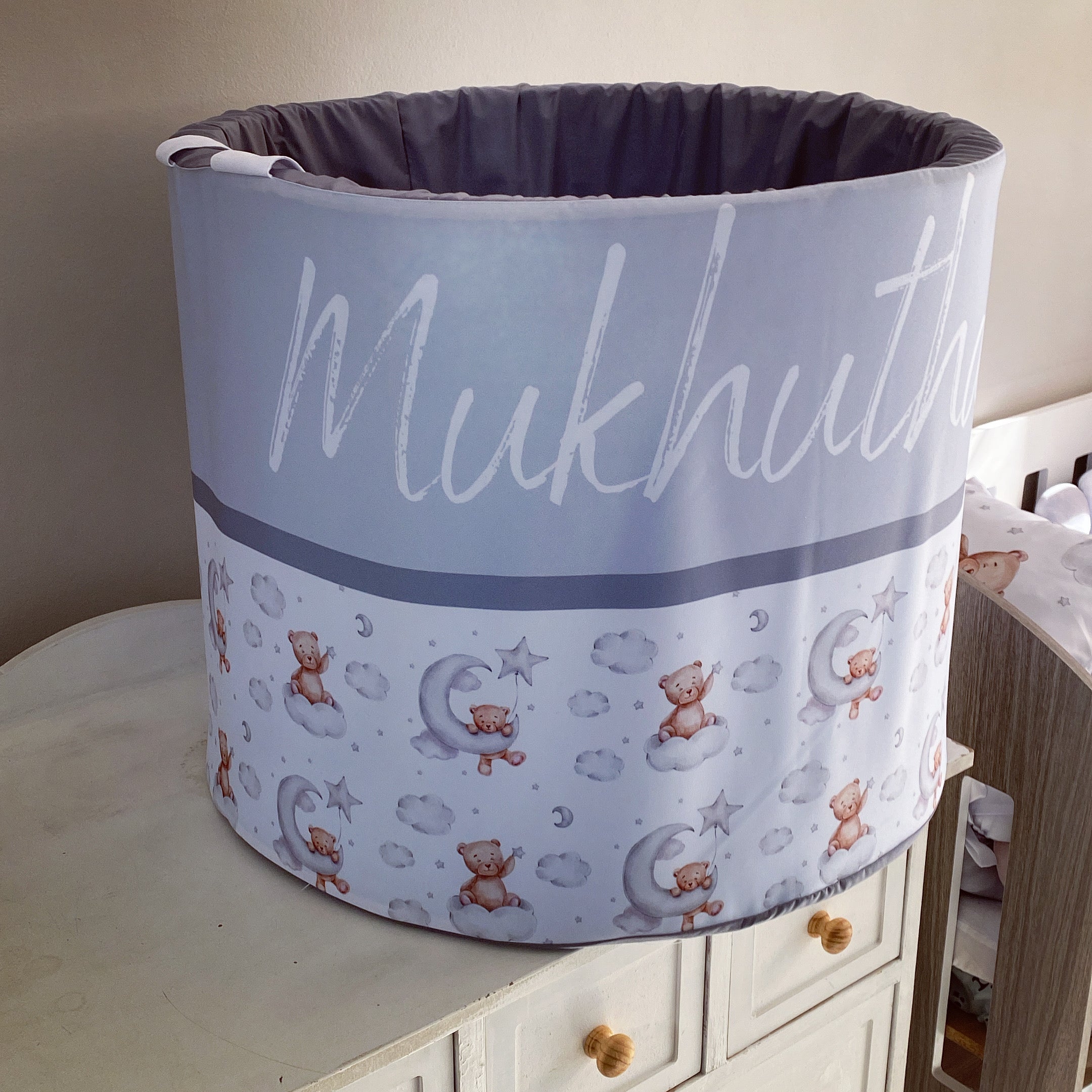 A large grey laundry barrel with a bear pattern and the text 'MUKHUTA' on it, placed on a wooden furniture item in a room with a light grey wall.