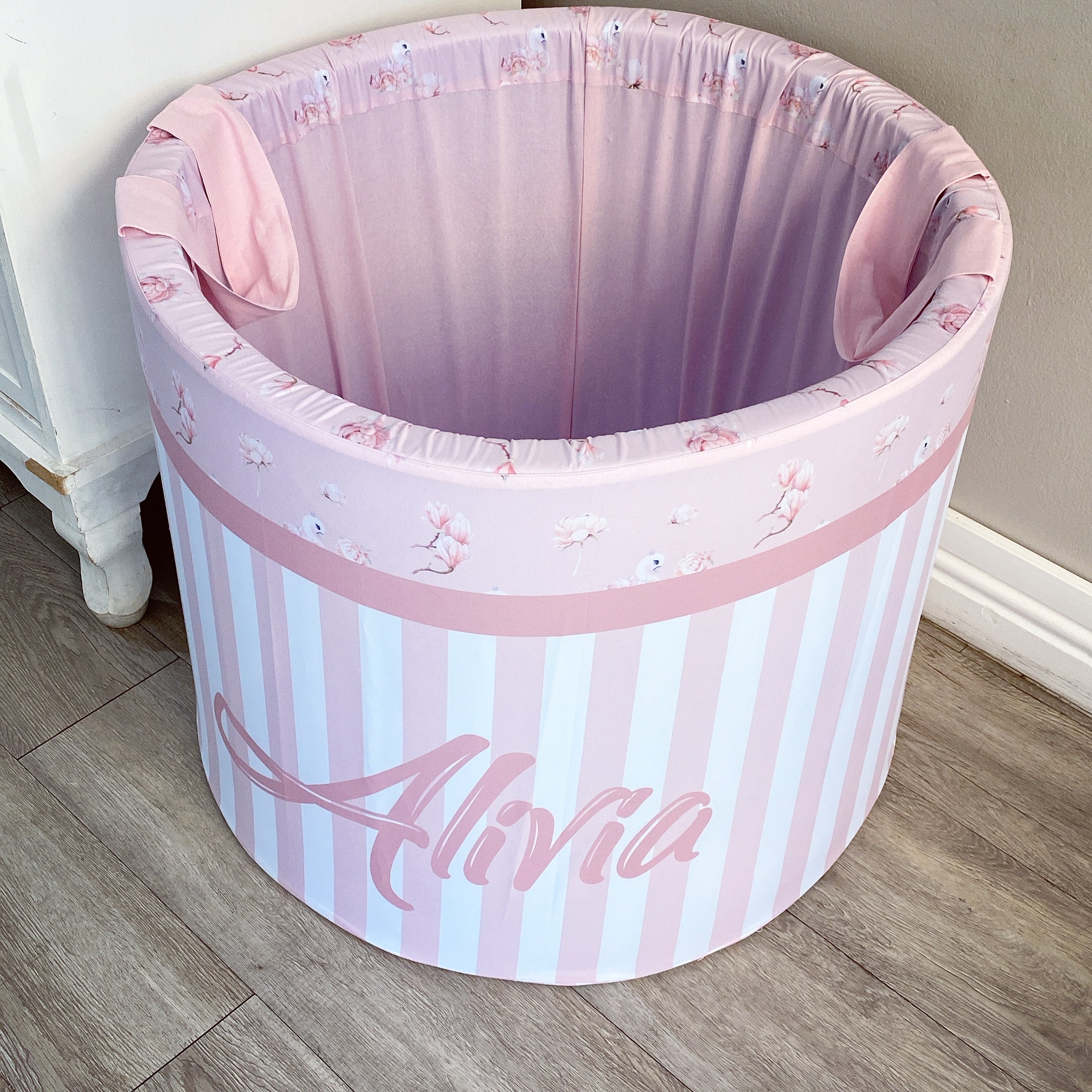 A large pink laundry barrel with striped patterns and floral designs, featuring the name 'Alva' on the side, placed in a room with light striped walls.