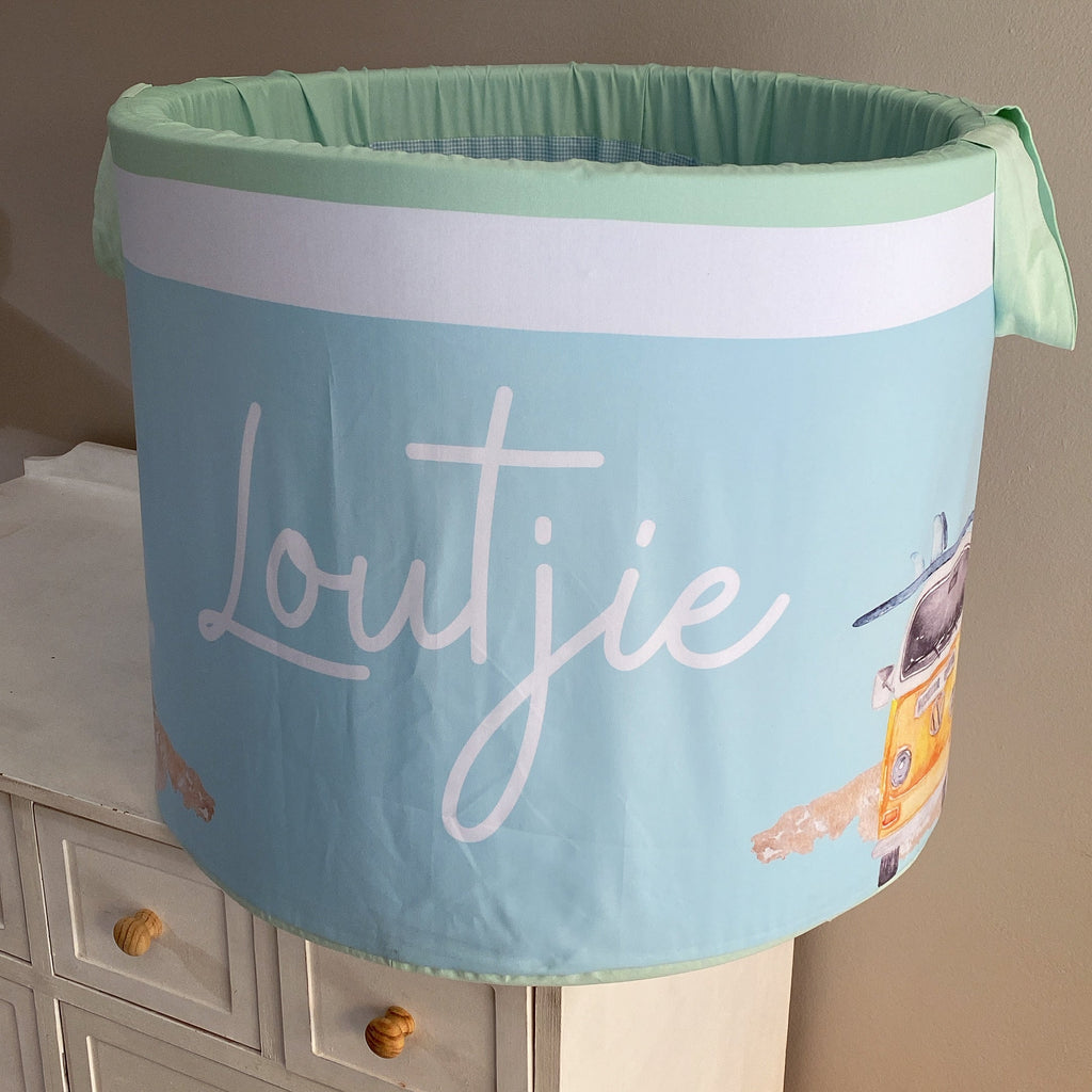 A large laundry basket with a green lid and white fabric, featuring the word 'LOUTJIE' in a large, cursive font, along with ocean-themed illustrations such as waves and seagulls.
