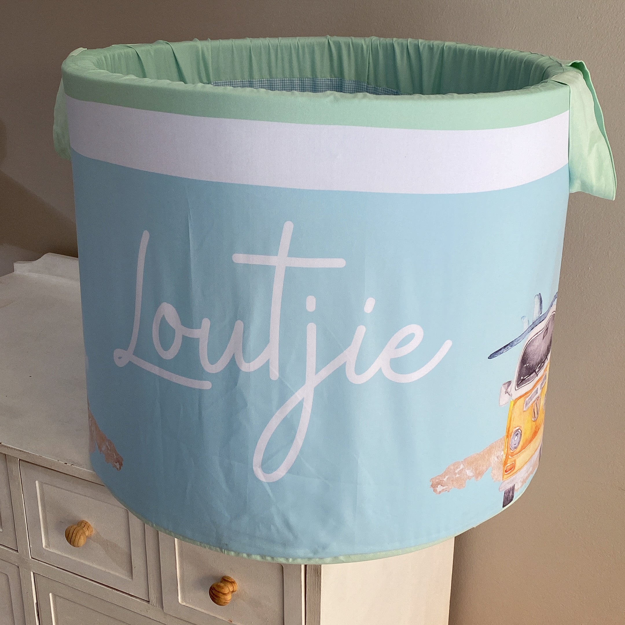A large laundry basket with a green lid and white fabric, featuring the word 'LOUTJIE' in a large, cursive font, along with ocean-themed illustrations such as waves and seagulls.