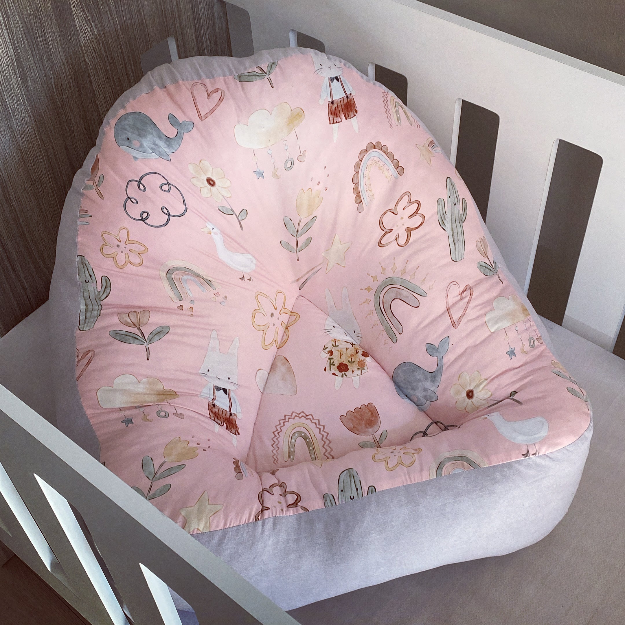 A pink boho-style baby lounger with a pattern of animals and plants, placed in a wooden crib.