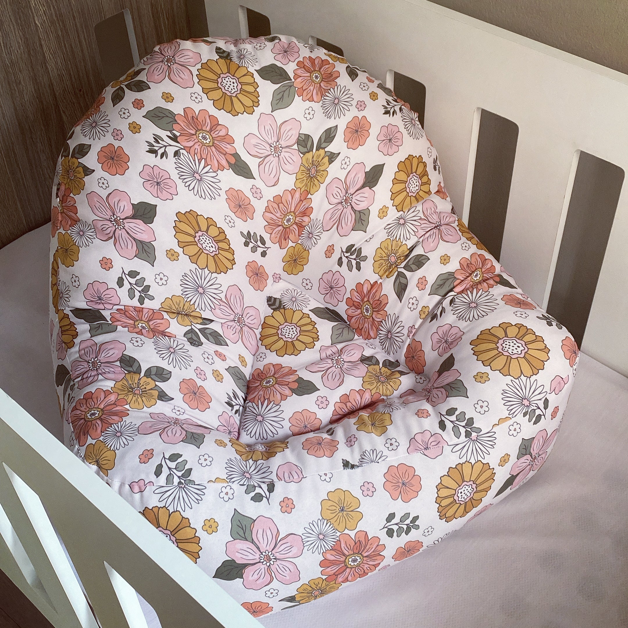 A floral-patterned lounger for babies, with a mix of pink and orange flowers on a white background, placed in a white crib.