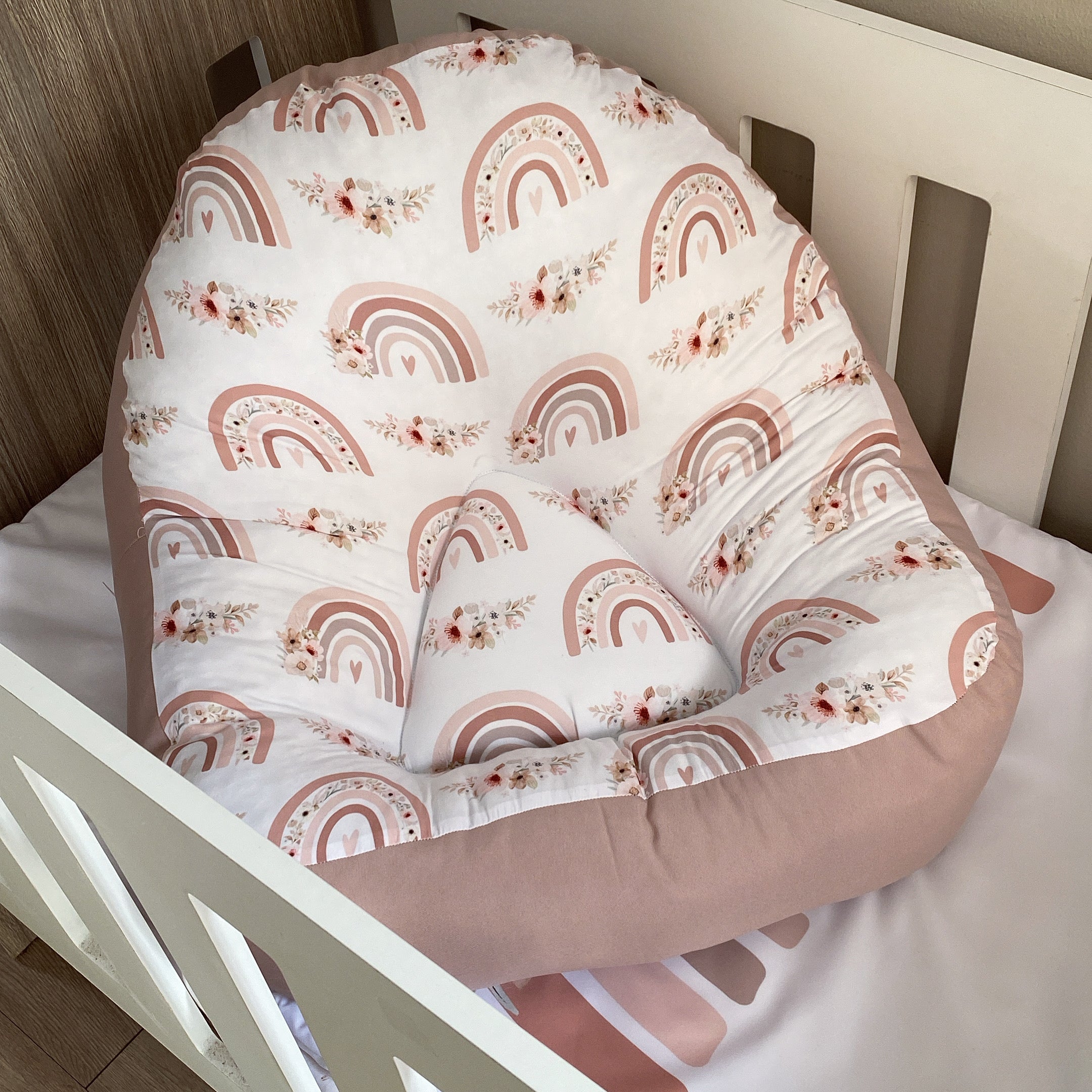A boho-style baby lounger with a white fabric featuring a rainbow and floral pattern, placed in a white crib.