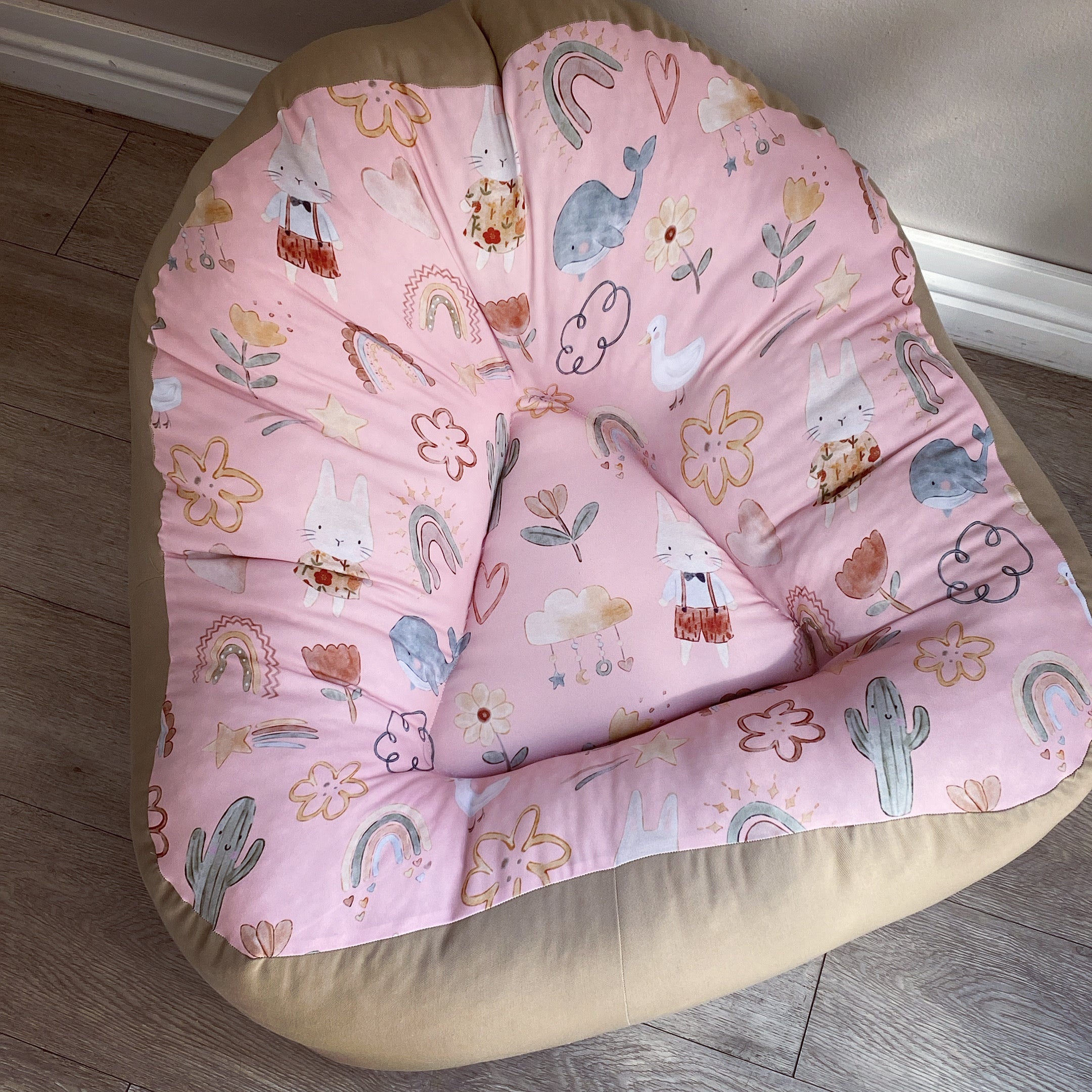 A pink bean bag chair with a pattern that includes bunny faces, cacti, and other playful elements, placed in a room with a wooden floor.
