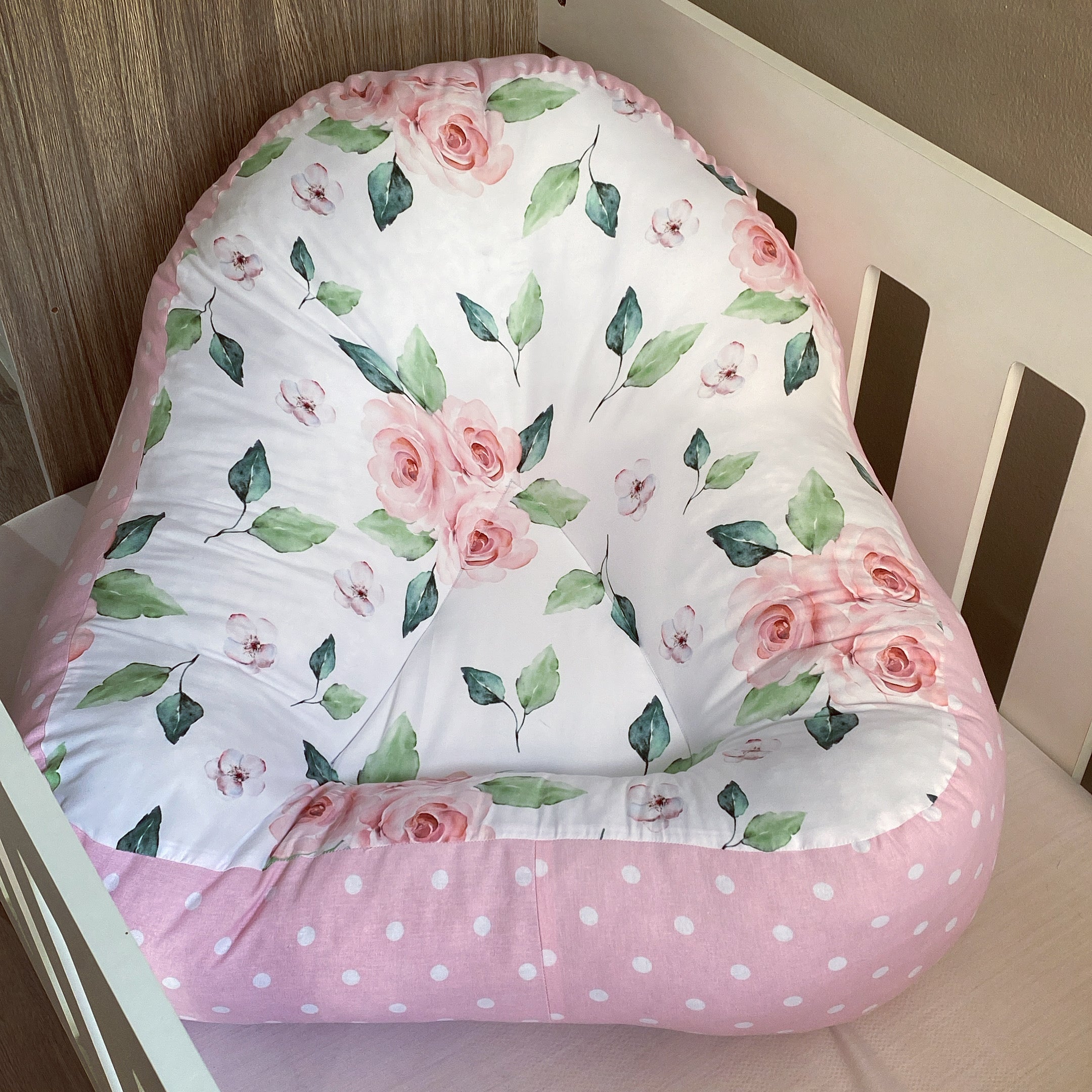 A pink floral-patterned baby lounger with a white wooden frame.