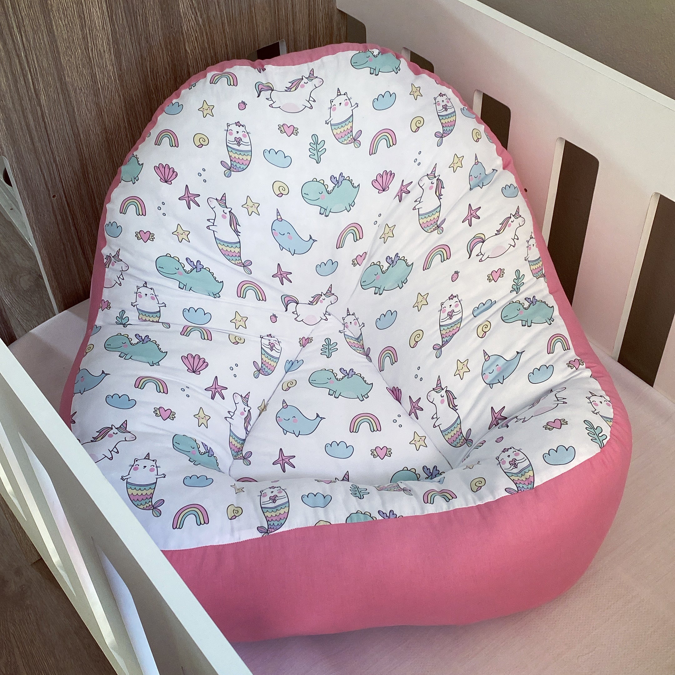 A pink fabric lounger with a unicorn and rainbow pattern, designed for toddlers, placed in a white crib.