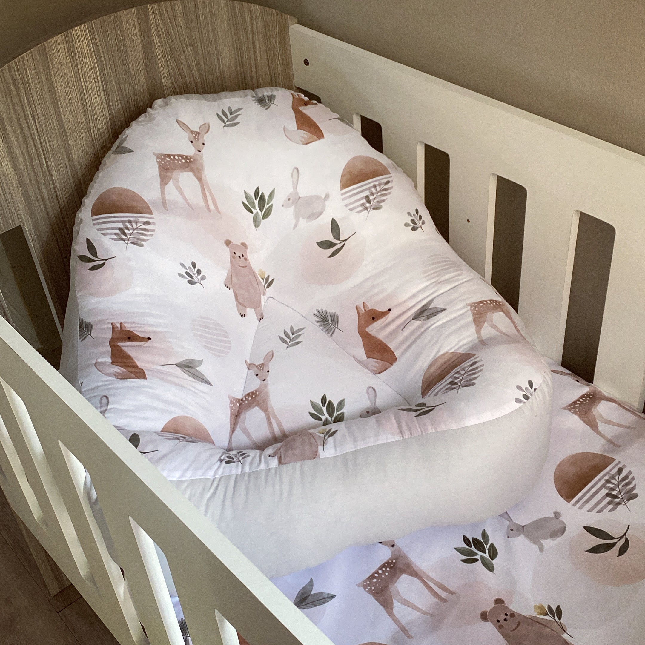 A white fabric lounger with a woodland animal print, positioned in a wooden crib.