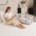 Woman reading a book next to a baby in a bassinet on a bed.