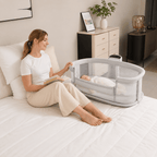 Woman reading a book next to a baby in a bassinet on a bed.