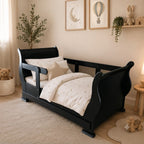 A black wooden children's bed with a curved headboard and footboard, styled with cream bedding in a warm, neutral-toned nursery room.