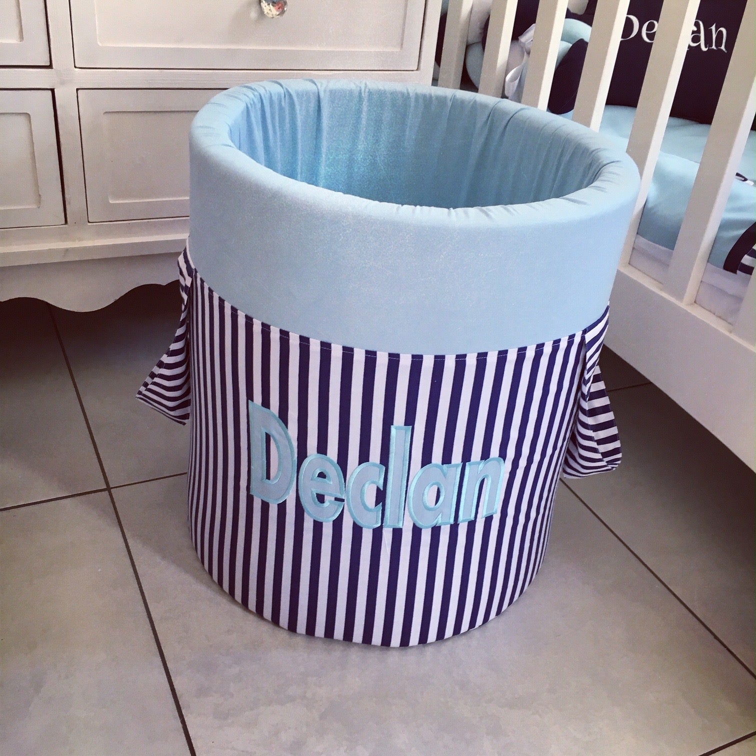 Striped laundry basket with blue interior and 'Declan' branding on a tiled floor.