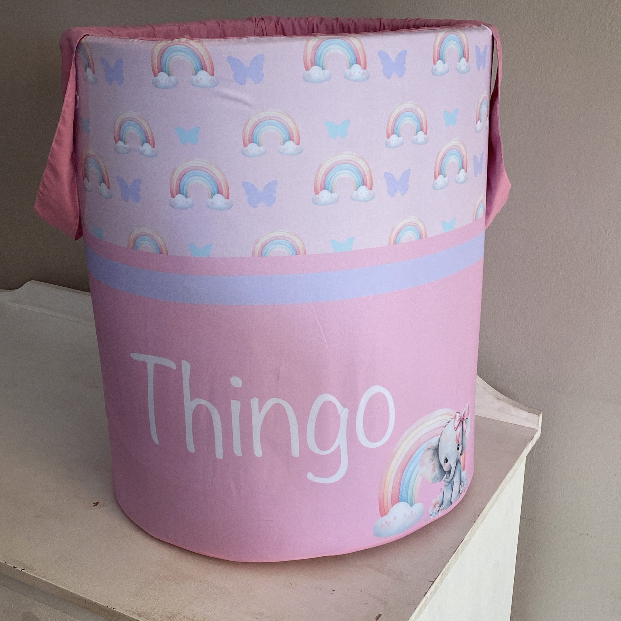 A pink laundry basket with a pattern of rainbows, clouds, and an elephant, and the word 'THINGO' printed on it.