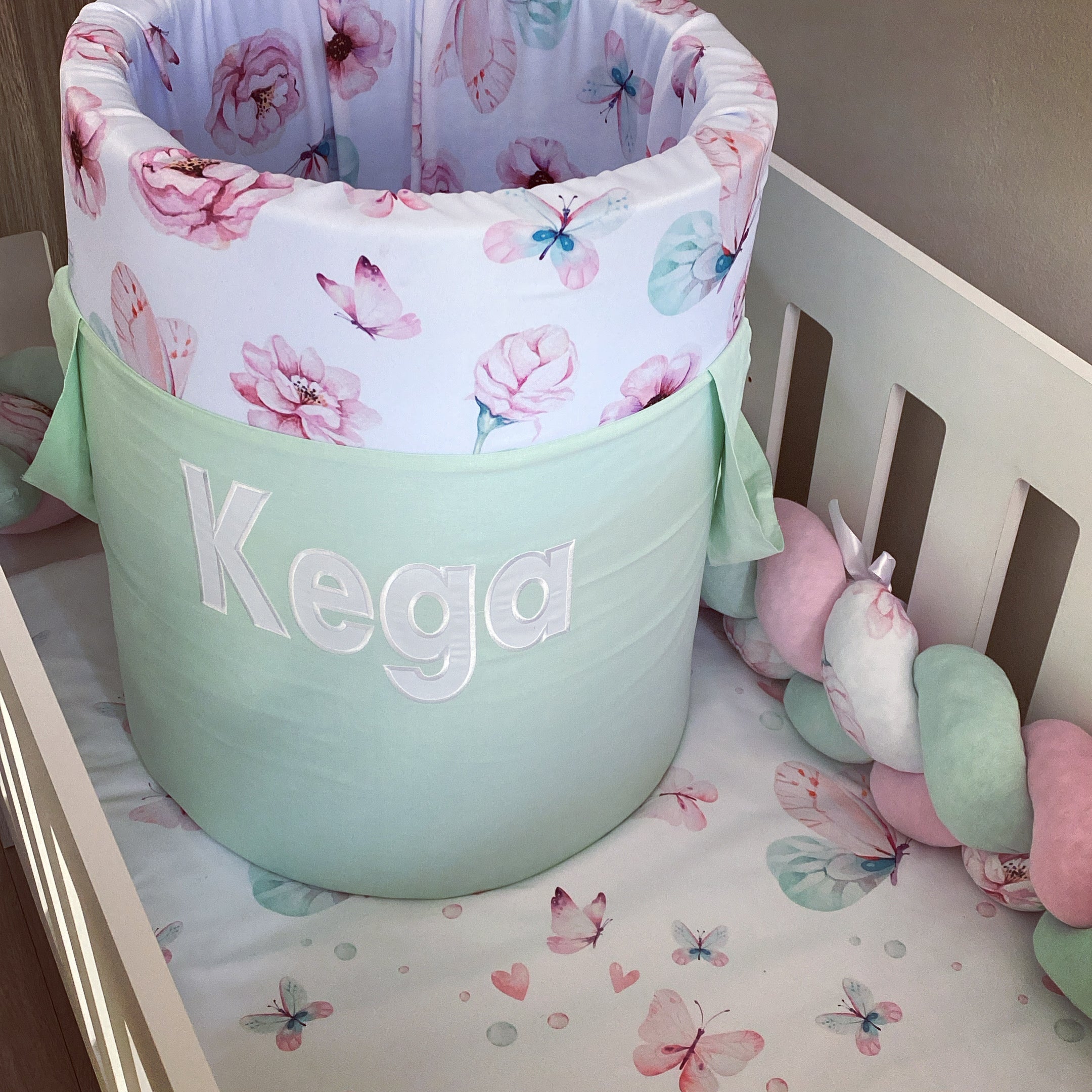 A mint-colored laundry basket with a floral pattern, including flowers and the name 'Kega' printed on it, placed in a nursery room setting with a few toys around.