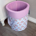 A pink laundry basket with a floral pattern and a pink lid, placed on a floor next to a white object, presumably a piece of furniture.