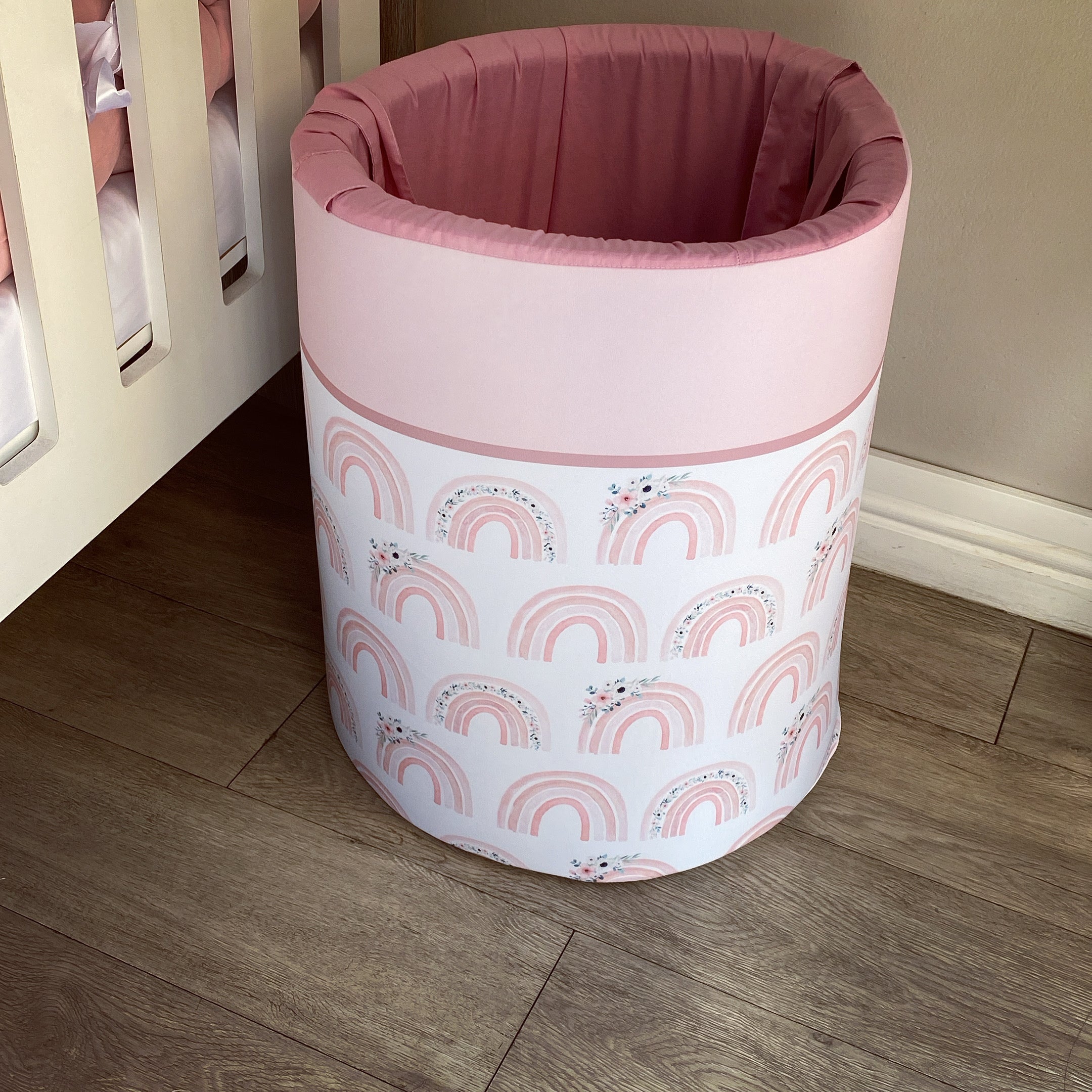 A pink laundry barrel with a rainbow and unicorn pattern, placed in a room corner with a wooden floor and a pink wall.