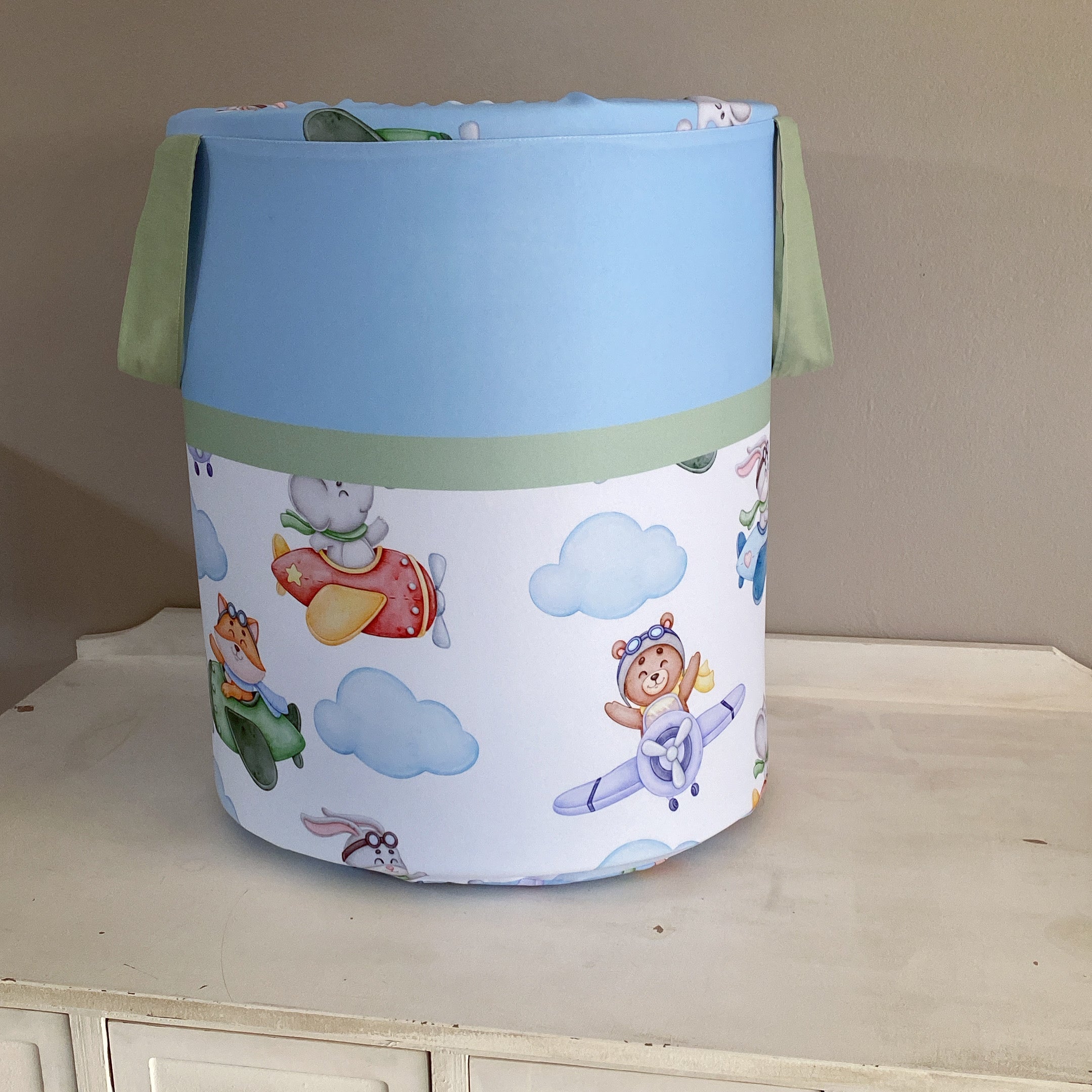 A blue laundry basket with a green lid, decorated with a pattern of airplanes, clouds, and cartoon characters.