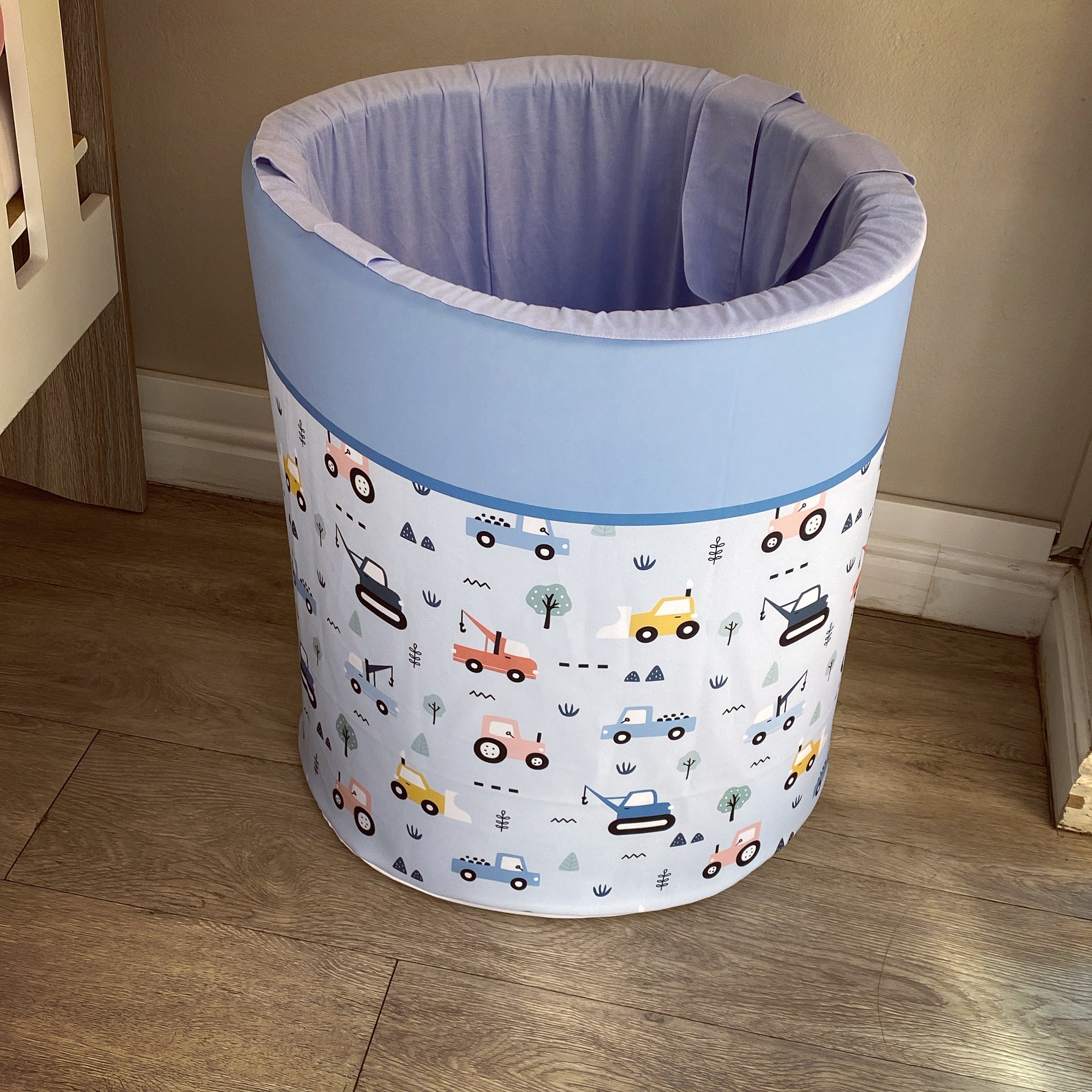 A blue laundry basket with a transportation theme, including cars and trucks, placed on a wooden floor.
