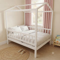 A white wooden house-shaped canopy bed with a slatted guard rail, styled with white and light gray patterned bedding, pillows, and a matching duvet in a child's bedroom with pink curtains.