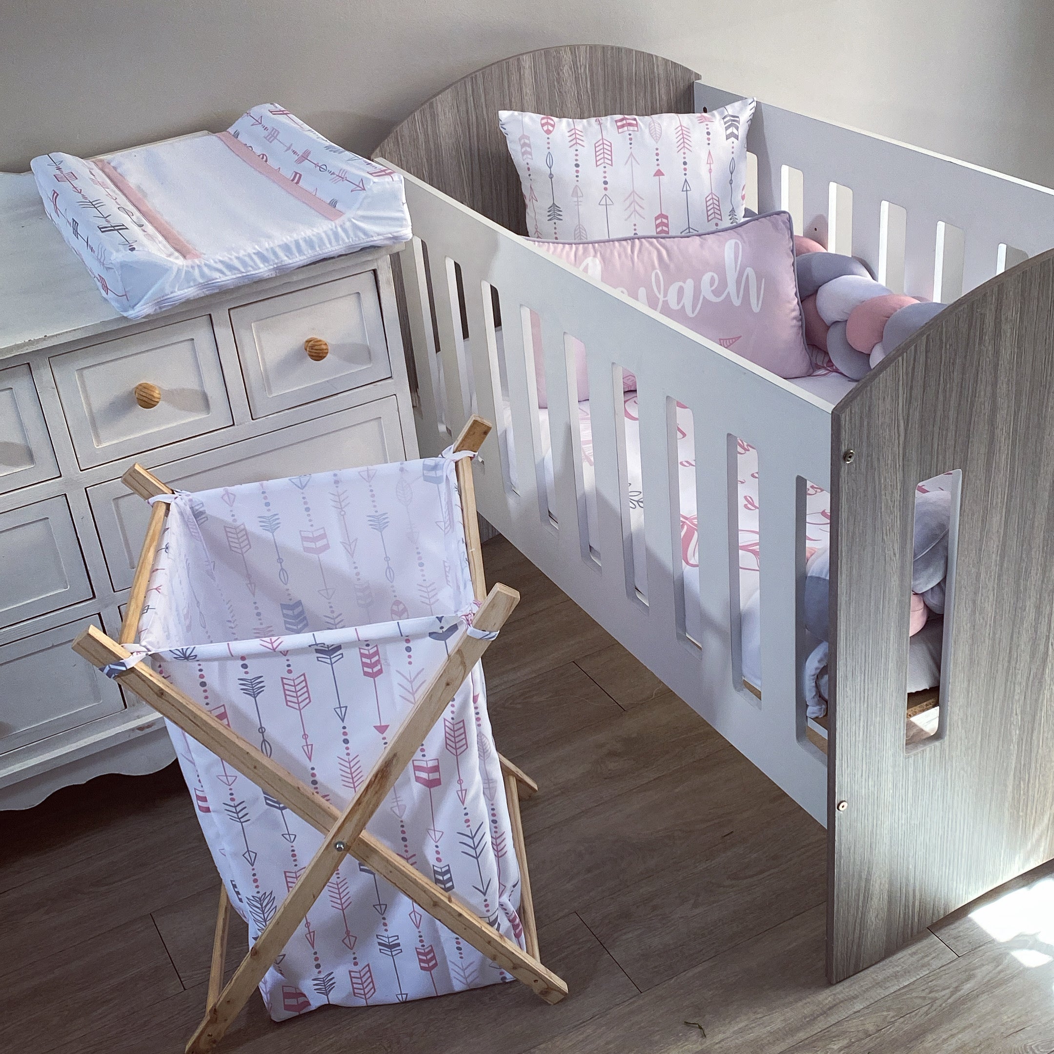 A wooden laundry basket with a white cushion and pink patterned fabric, placed in a nursery room setting with a wooden crib and a side table.