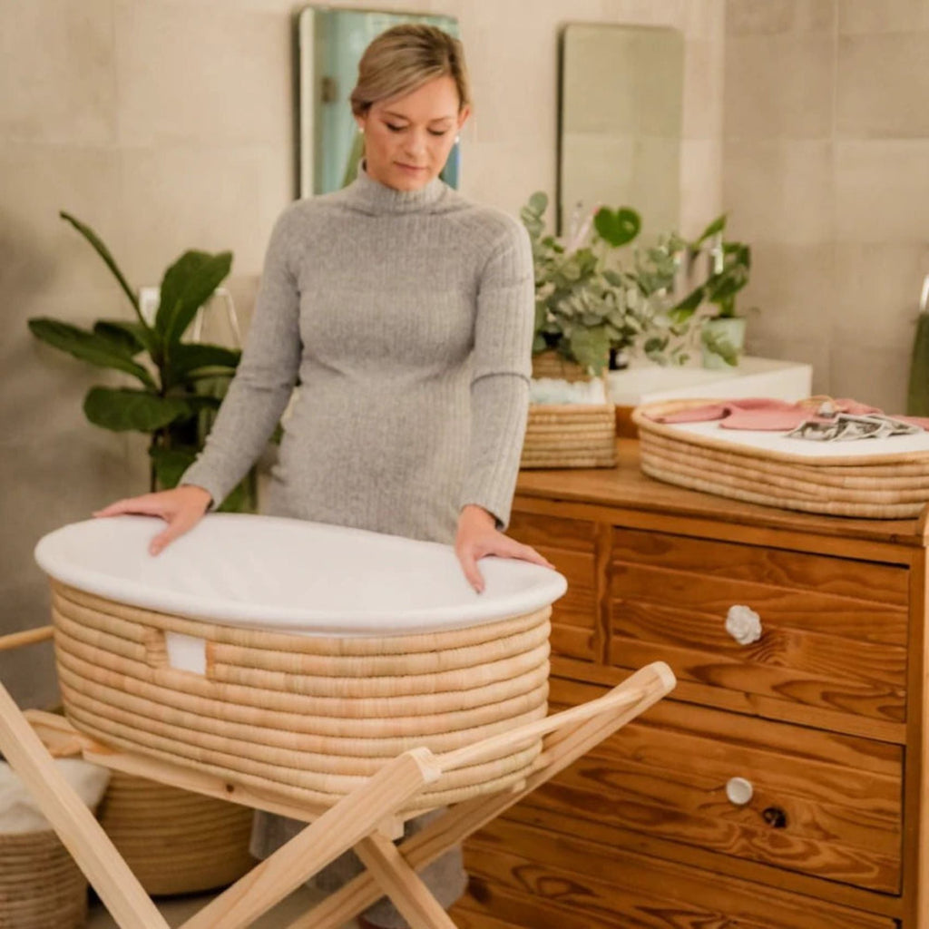Woman holding a woven baby crib in a room with wooden furniture and plants.