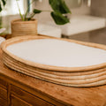 Woven basket with a white cushion on a wooden surface, with a blurred background of a plant and furniture.