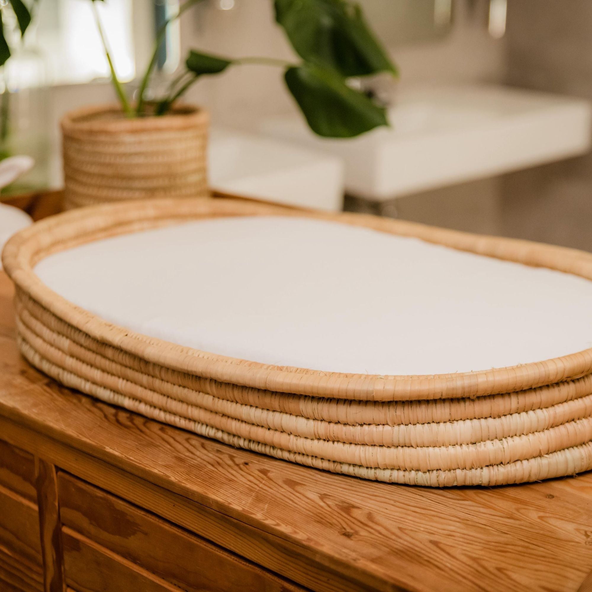 Woven basket with a white cushion on a wooden surface, with a blurred background of a plant and furniture.