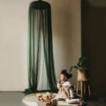 A child sitting under a green emerald hanging tent canopy netting, with the canopy casting a shadow on the wall behind.