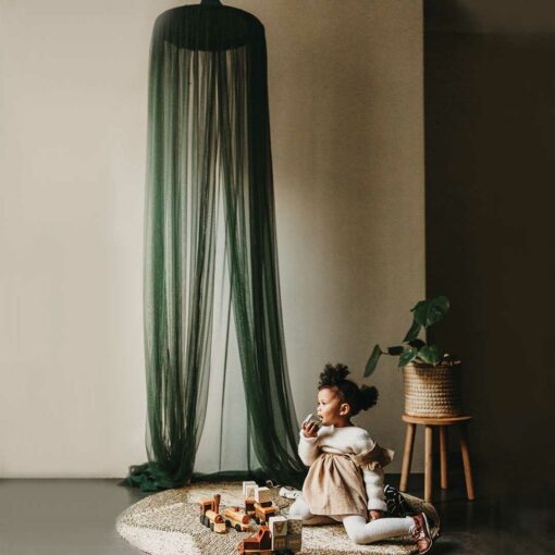 A child sitting under a green emerald hanging tent canopy netting, with the canopy casting a shadow on the wall behind.